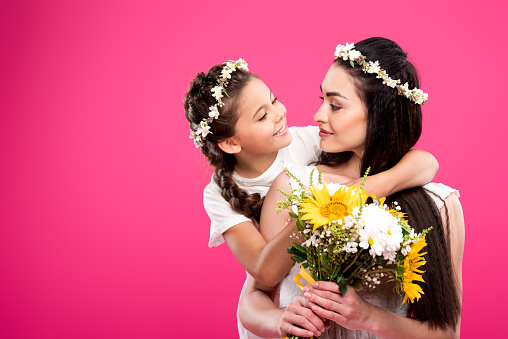 adorable happy daughter looking at beautiful mother holding flower bouquet isolated on pink