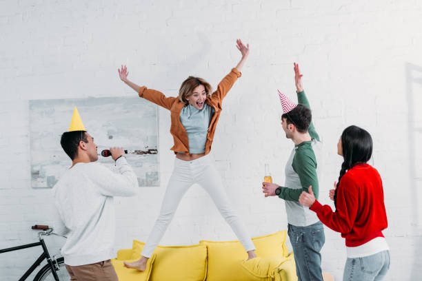 istock cheerfull girl jumping on yellow sofa while multicultural friends singing karaoke and dancing 1151139976