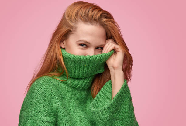 Adorable flirty girl hiding face in sweater Young redhead woman hiding face in neck of warm green sweater looking at camera on pink backdrop shy stock pictures, royalty-free photos & images