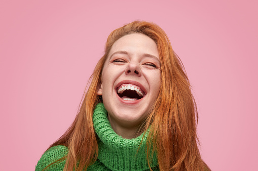 Contemporary vivid redhead woman in green sweater laughing emotionally on pink backdrop