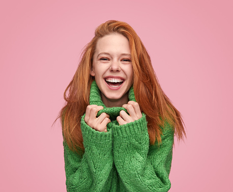 Modern ginger woman cuddling in cozy green sweater and laughing at camera on pink background