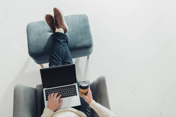 cropped view of man holding paper cup and using laptop with blank screen