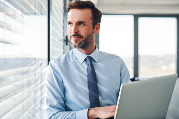 Handsome businessman standing with laptop, looking through the window in modern office