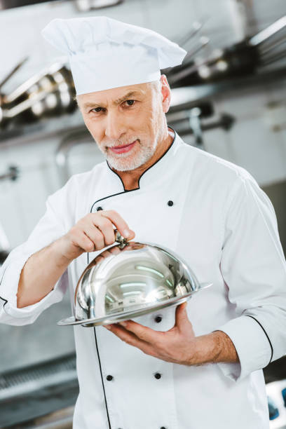 istock handsome male chef in uniform looking at camera and holding serving tray with dome in restaurant kitchen 1151157433