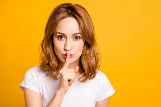 Close up photo beautiful amazing she her lady hold arm hand index finger mouth lips ask stop talking tell speak say secrecy information wear casual white t-shirt isolated yellow background.
