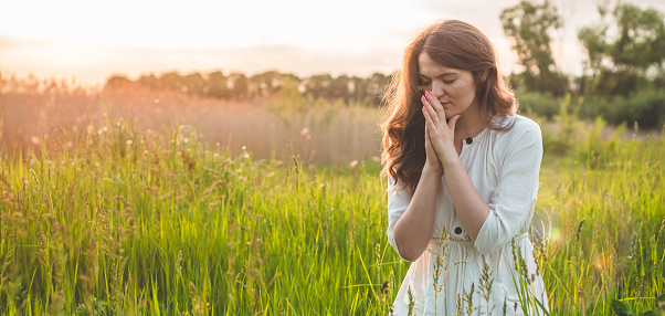 Girl closed her eyes, praying in a field during beautiful sunset. Hands folded in prayer concept for faith, spirituality and religion. Peace, hope, dreams concept