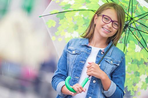 Portrait of beautiful young pre-teen girl with umbrella under spring or summer rain. Smilling girl with dental braces and glasses.