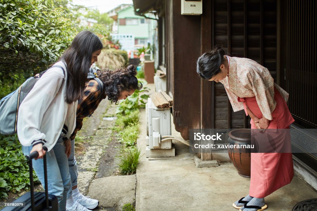 Landlady greeting female guests outside ryokan Mature landlady greeting young female guests at ryokan. Women are bowing in front of inn owner. They are standing outside hotel. Japan Stock Photo
