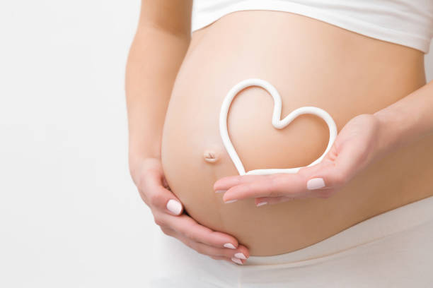 die hand der jungen frau hält weiße herzform auf nacktem bauch. emotional liebevoller moment in der schwangerschaftszeit-30 wochen. baby-erwartung. liebe, glück und sicherheitskonzept. closeup. - human pregnancy abdomen naked human hand stock-fotos und bilder