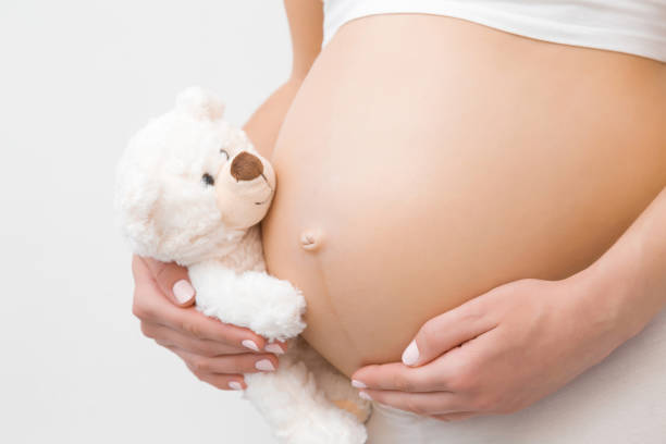 hand hält lächelnden weißen teddybären. nackte bauch der jungen frau. emotional liebevoller moment in der schwangerschaftszeit-30 wochen. baby-erwartung. liebe, glück und sicherheitskonzept. closeup. seitenansicht. - baby beauty beautiful the human body stock-fotos und bilder