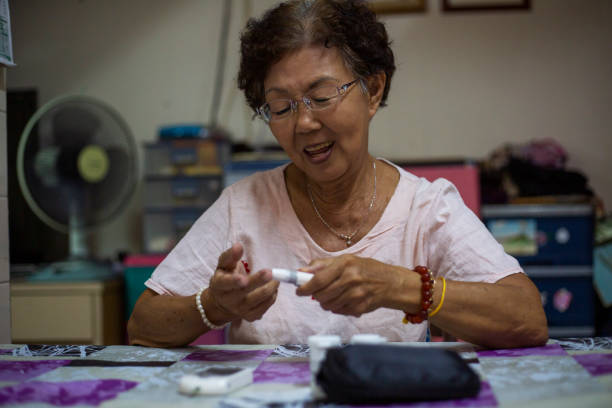 ältere asiatische diabetikerin, die ihren blutzucker mit einem glaukometer überprüft - glaucometer analyzing blood equipment stock-fotos und bilder