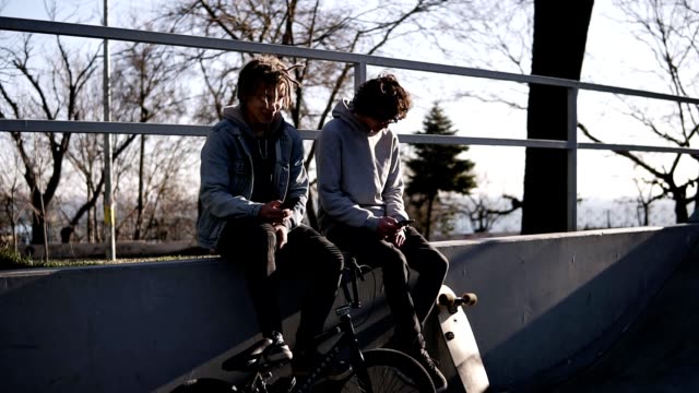 Two friends skateboarder and bmx rider are sitting together on high parapet in the city skate park using their smartphones. Texting, communication. Sunny day. Active leisure time, sport and technology concept