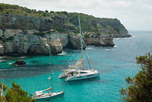 Catamaran and sailing boat in the turquoise sea of the spanish island with high cliff as panorama