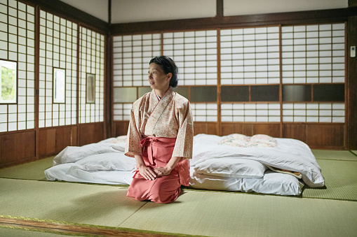 Thoughtful landlady of ryokan kneeling on tatami mat. Female owner is looking away. She is in kimono.