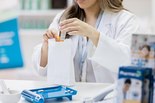 Unrecognizable female pharmacist places a customer's prescription medication into a paper bag. A mortar and pestle and pill counter are on the counter in front of her.