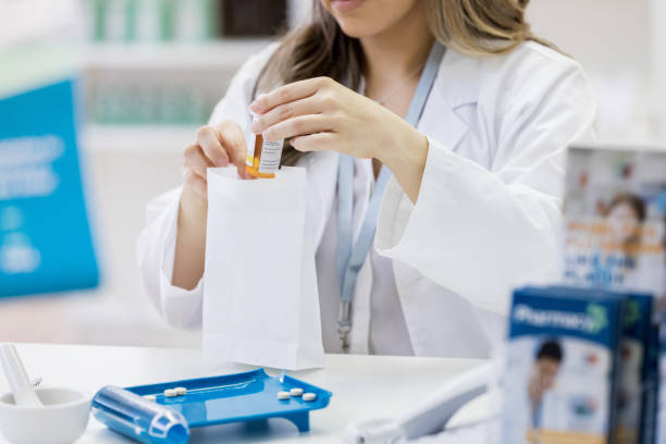 Pharmacist bags up a customer's prescription Unrecognizable female pharmacist places a customer's prescription medication into a paper bag. A mortar and pestle and pill counter are on the counter in front of her. pharmaceutical compounding stock pictures, royalty-free photos & images