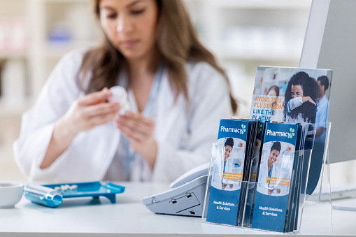 A pharmacist concentrates while filling a patient's prescription. She is holding a pill bottle. A pill counter is in front of her. Focus in on informational brochures in the foreground.