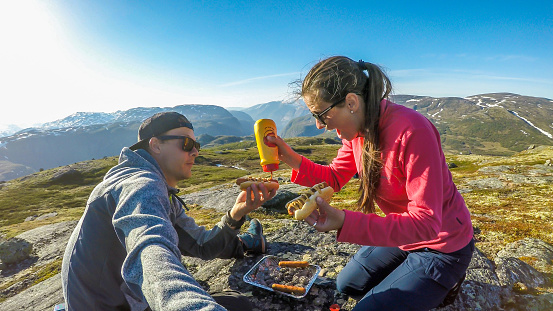 A couple having a grill in the tall mountains. Few sausages are being grilled. Couple uses portable one time grill, picnic grill. Girl spreads the mustard over the hot dogs. Camping in wilderness.