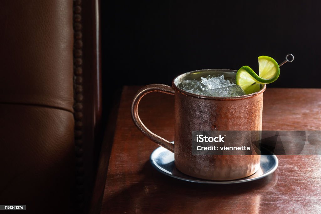 Moscow Mule Cocktail in Copper Mug in Dark Luxurious Bar with Copy Space A Moscow Mule cocktail in a copper mug with crushed ice and a lime garnish on a dark wood table next to a leather seat. This drink is made with vodka, ginger beer (or ginger ale), and lime juice. Taken in a dark luxurious bar or restaurant. Cocktail Stock Photo