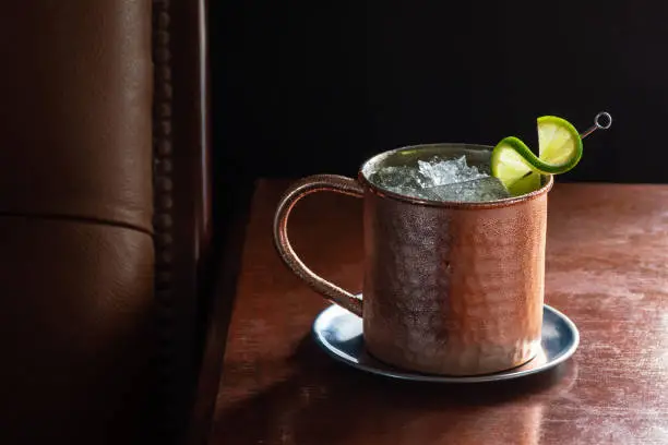 A Moscow Mule cocktail in a copper mug with crushed ice and a lime garnish on a dark wood table next to a leather seat. This drink is made with vodka, ginger beer (or ginger ale), and lime juice. Taken in a dark luxurious bar or restaurant.
