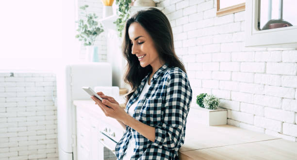 hermosa mujer sonriente linda está utilizando el teléfono inteligente en la cocina en casa. - on the phone women beautiful beauty fotografías e imágenes de stock