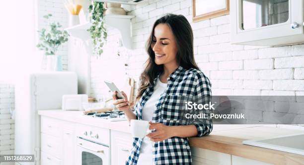 Beautiful Cute Smiling Woman Is Using Smart Phone On The Kitchen At Home Stock Photo - Download Image Now
