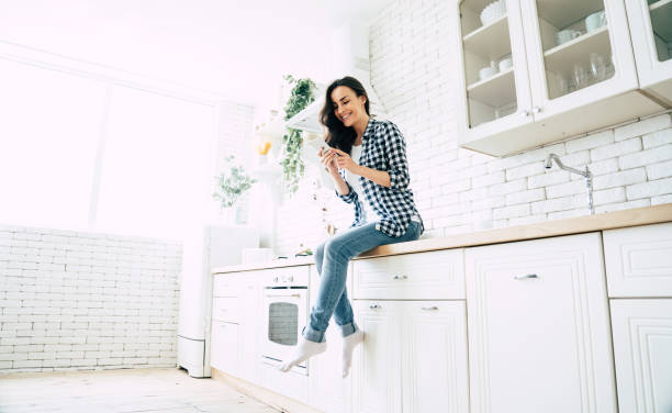 Beautiful cute smiling woman is using smart phone on the kitchen at home.