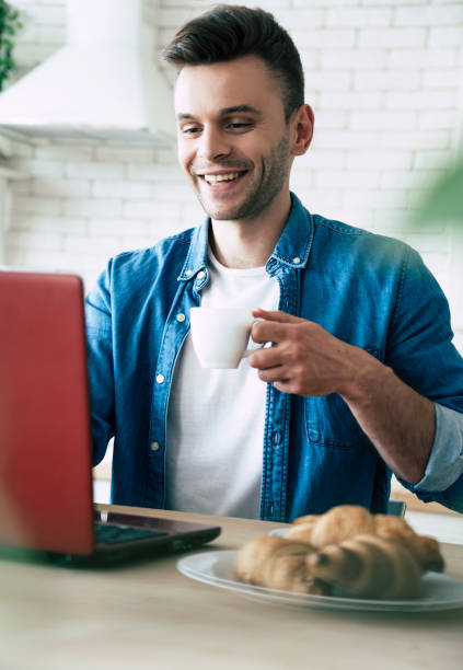 istock Happy morning. Beautiful smiling man is using laptop and drinks coffee or tea while sitting on the kitchen at home 1151268626