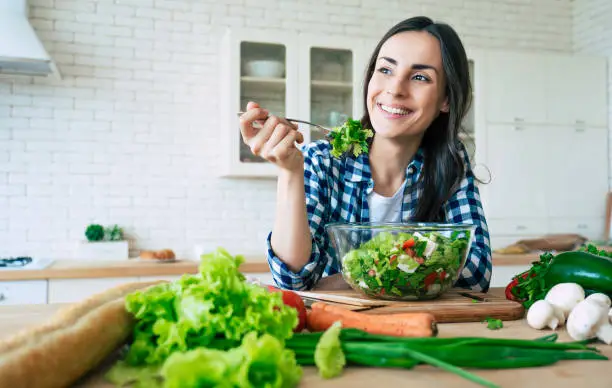 Photo of Healthy lifestyle. Good life. Organic food. Vegetables. Close up portrait of happy cute beautiful young woman while she try tasty vegan salad in the kitchen at home.