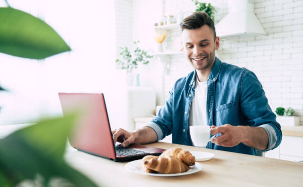 istock Happy morning. Beautiful smiling man is using laptop and drinks coffee or tea while sitting on the kitchen at home 1151268644