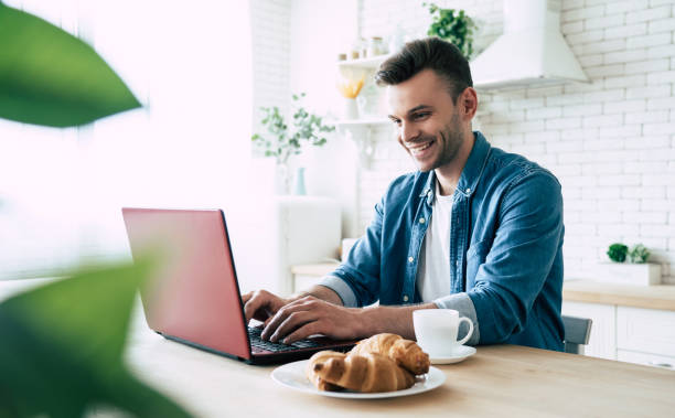 istock Happy morning. Beautiful smiling man is using laptop and drinks coffee or tea while sitting on the kitchen at home 1151268664