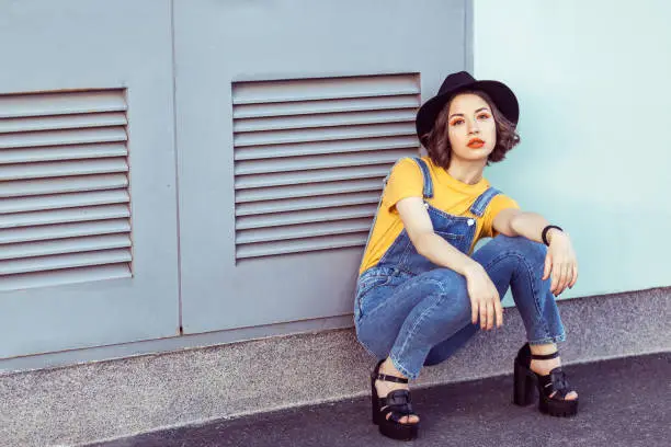 Photo of young woman in blue denim overalls and yellow tshirt with black hat sensual looking at camera while posing near industrial building