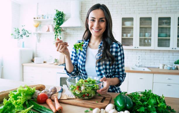 Healthy lifestyle. Good life. Organic food. Vegetables. Close up portrait of happy cute beautiful young woman while she try tasty vegan salad in the kitchen at home. stock photo