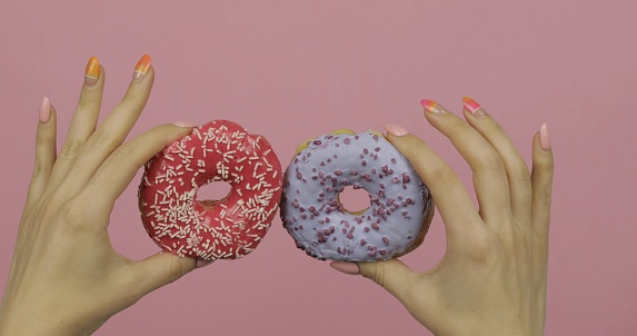Womens hands holding two red and blue, delicious, tasty and fresh donuts on pink background. Bright and colorful sprinkled sweet donut in girl hand
