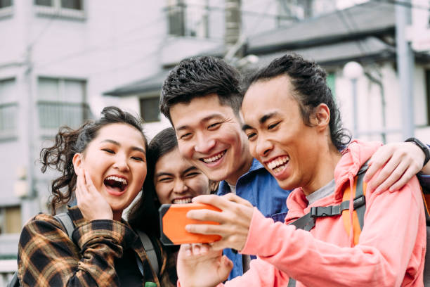 Group of four cheerful friends looking at smartphone and laughing Multi racial young adults using mobile phone, arm around, joy, social media, communication central asian ethnicity stock pictures, royalty-free photos & images