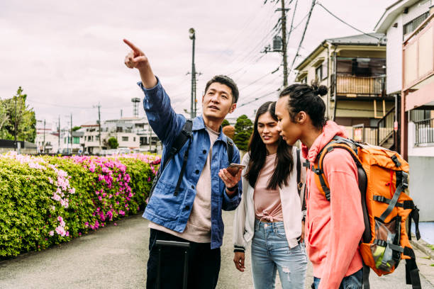 Young man with smartphone pointing and talking to friends with backpack Three young adults travelling in city, using GPS on phone, arriving, destination, location central asian ethnicity stock pictures, royalty-free photos & images