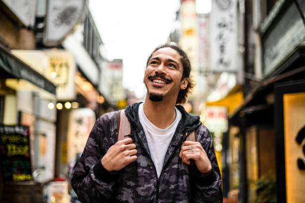 Cheerful young man with goatee looking up in city street Male traveller carrying backpack exploring new city and smiling, travelling alone, discovery, adventure, independence male meaning stock pictures, royalty-free photos & images