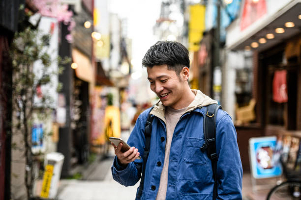 Cheerful young man looking at smartphone in street Young Chinese man in his 20s smiling and checking mobile phone, carrying backpack, city life, communication, social media asian tourist stock pictures, royalty-free photos & images