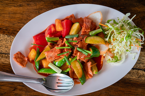 Roasted Pork with vegetables: tomato, pepper, green onion, cabbage with sauce in white plate, on wooden table.