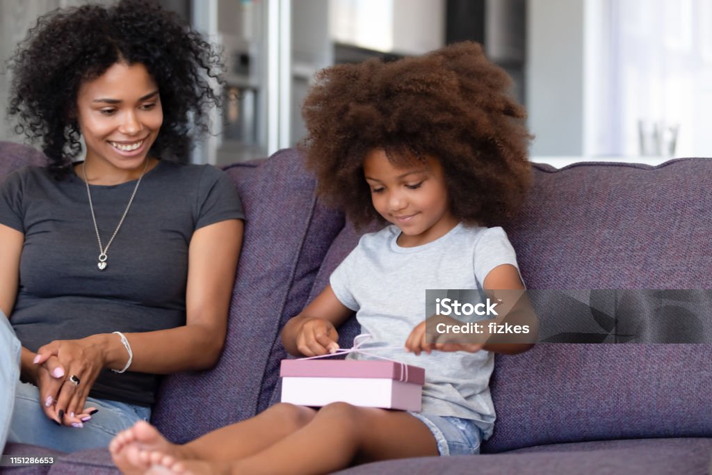 Cute little black girl unwrap birthday present at home Happy young African American mom watch small cute daughter unpack birthday gift sitting on couch together, little black girl unwrap package presented by mother on special occasion at home Birthday Present Stock Photo