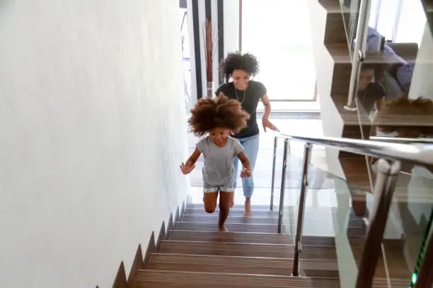 Excited little African American girl run up stairs of new home with young mom, happy black mother and small daughter explore bought house on moving day together, mommy have fun playing with kid