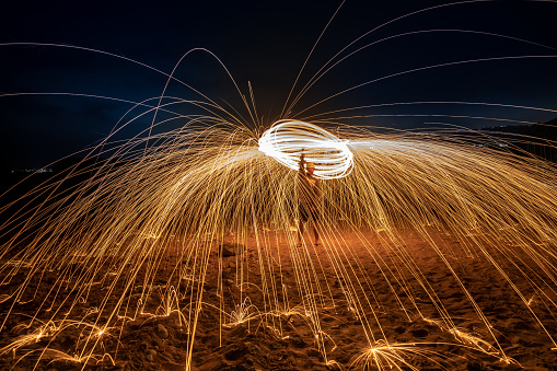 A young adult male spins flaming steel wool as the camera captures the sparks bouncing off the sand with a long exposure.