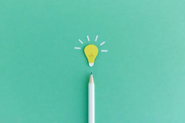 Creative composition of pencil with small paper light bulb glowing above on green background