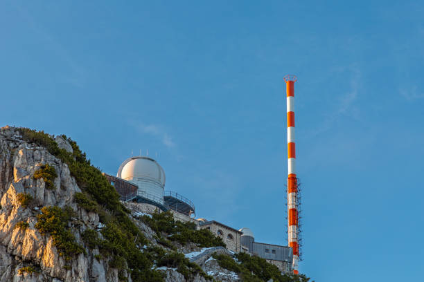 Metal pyramid summit marker on rocky mountain top surrounded by communication antennas. Outdoor mountain peak with panoramic sky and distant mountain views. High altitude landscape and technology.