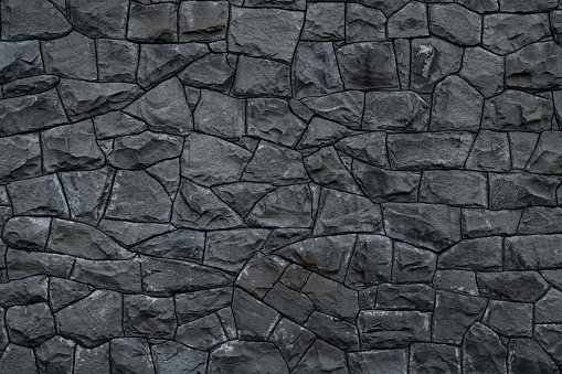 Gray dirty stone wall. Texture of grey granite. Dark rough rocks background. Weathered dark gray grunge building's facade. Stone surface. Mosaic pattern of grey stones on the cement wall. Old dirty wall rock texture. Grey backdrop of decorative tiles.