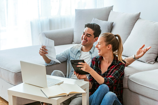 Photo of a happy couple doing finances at home. Photo of a cheerful loving young couple using a laptop and analyzing their finances with documents. Looking at papers. Happy couple at home paying bills with the laptop. Loving young couple using a laptop and analyzing their finances. Writing notes.