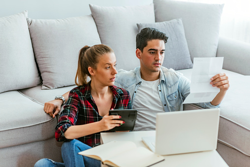 Photo of Depressed young couple doing their accounting in their living room during the day. Young couple calculating their domestic bills at home. Family budget and finances. Young woman doing accounts together with her husband at home, planning new purchase. Serious female making necessary calculations.