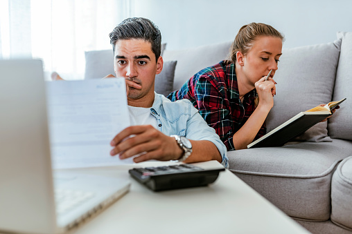 Young couple managing finances, reviewing their bank accounts using laptop computer and calculator at home. Woman and man doing paperwork together, paying taxes online on notebook pc. Photo of Depressed couple doing their accounting in their living room