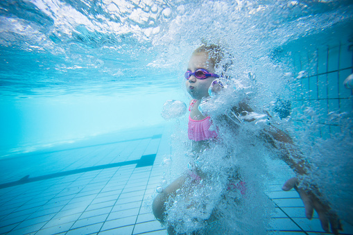 Wide angle underwater photo of a toddler girl swimming in a big swimming pool with goggles and a pink bikini