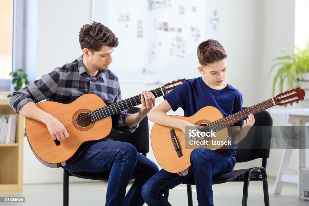 Guitarist teaching boy plucking string instrument Guitarist teaching boy plucking string instrument at classroom. Males are practicing guitar at conservatory. They are in training class. 12-13 Years Stock Photo